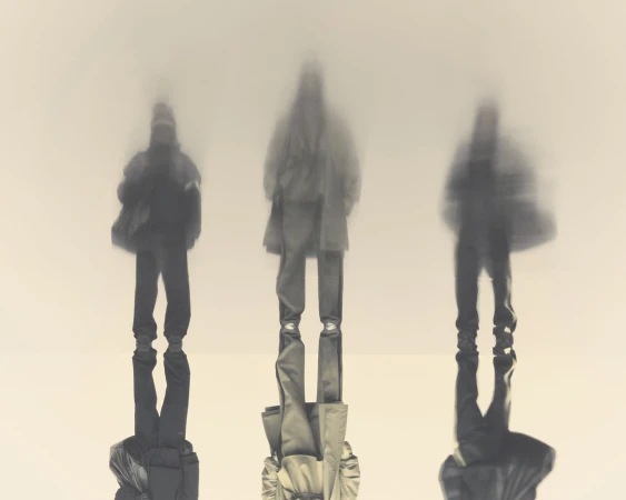Three blurred figures wearing coats and trousers, standing outdoors, abstract appearance, desaturated colour palette.