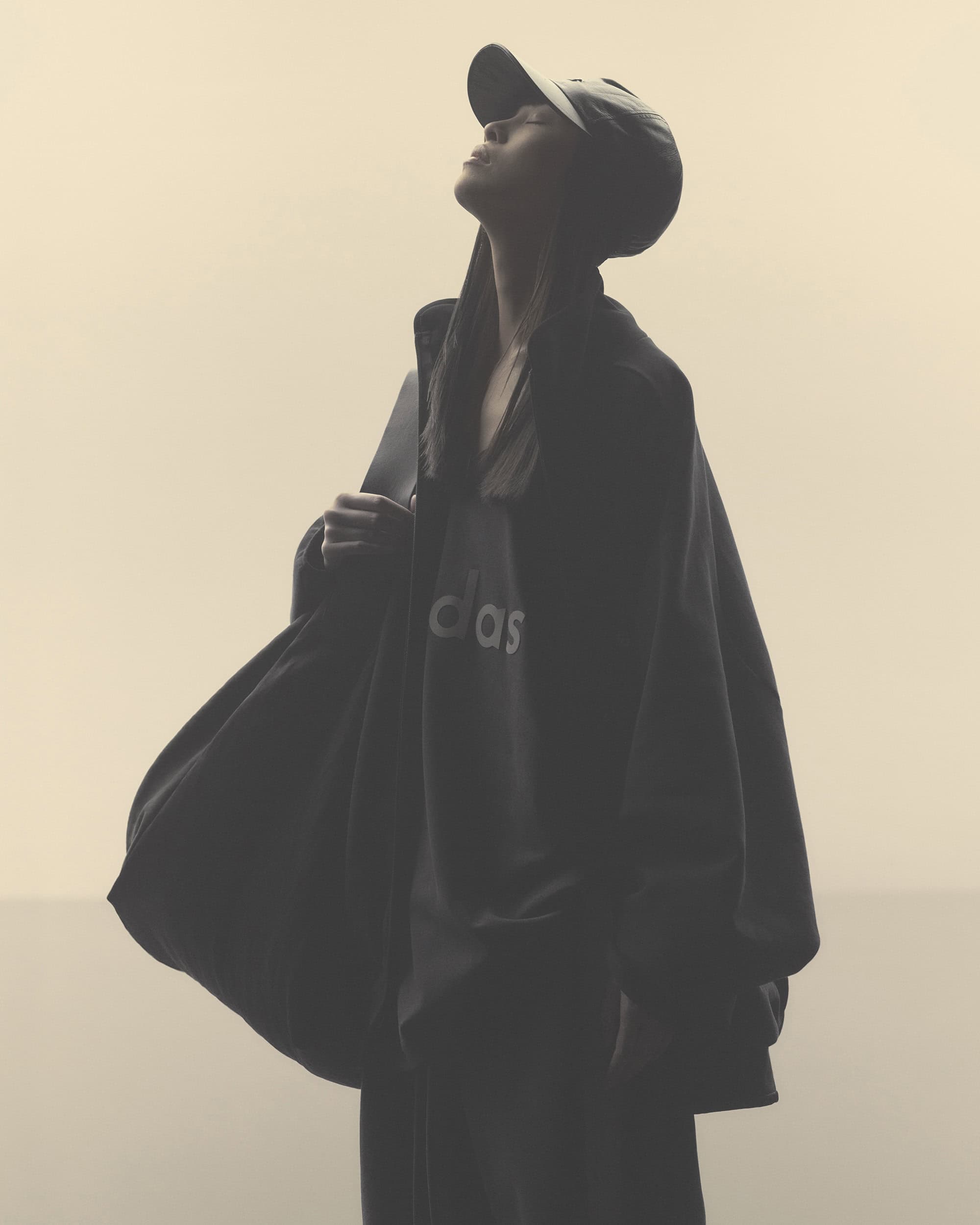 Adult female in Adidas hooded sweatshirt and baseball cap gazes upward in a muted, monochromatic outdoor fashion shot.