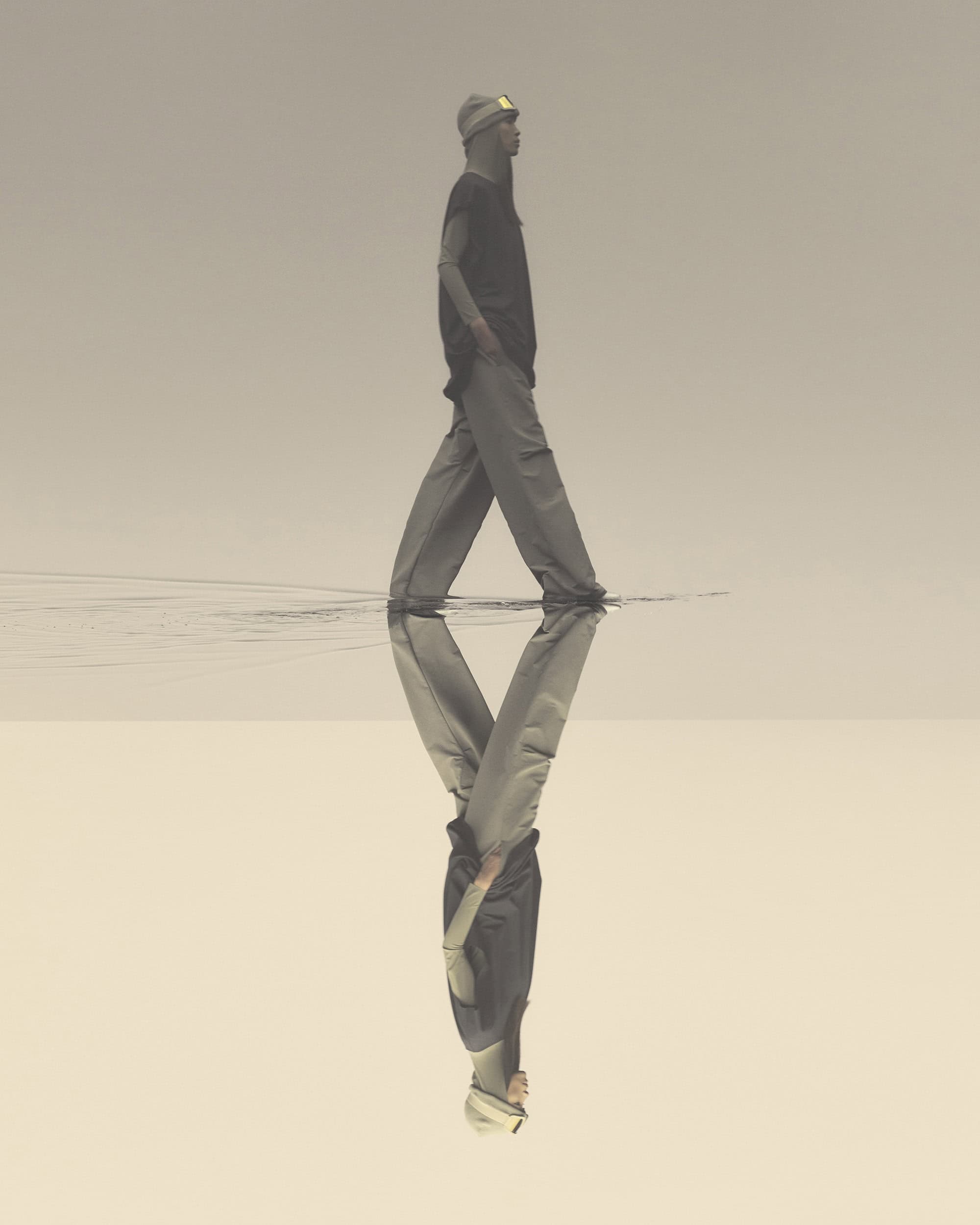 A man walks in shallow water, wearing casual clothes and a beanie, reflected in the water's surface against a neutral sky.