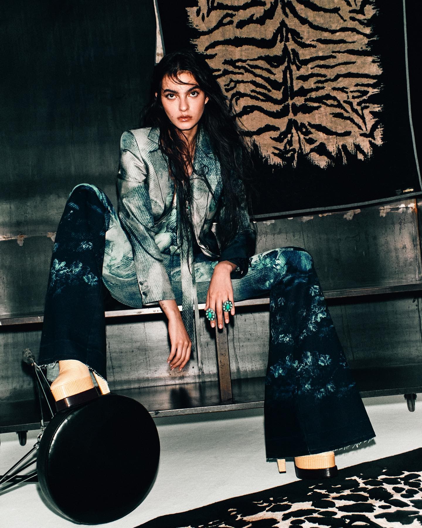Fashion shot of a brunette woman in patterned suit sitting on a metal bench, with a black drum and tiger-print textile backdrop.