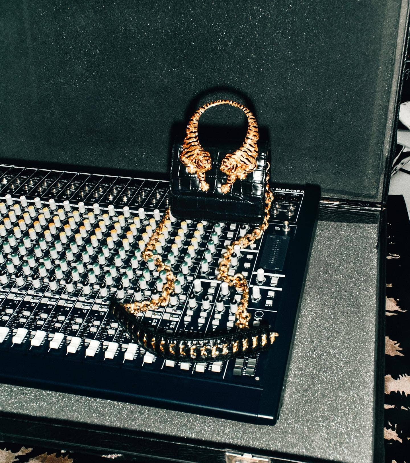 Close-up studio shot of a black purse with gold tiger heads on top of a sound mixing board inside of a black padded case.