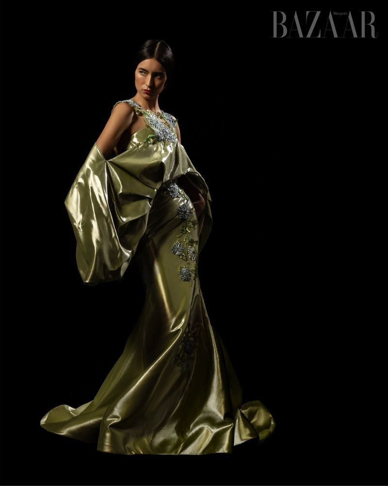 A woman poses in a full-length olive-green gown in a dramatic portrait with a black backdrop, showcasing the gown's shimmering fabric and floral embellishments.