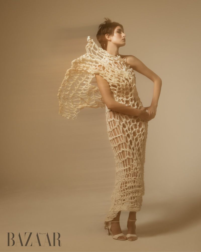 Fashion shot of a model wearing a long, knitted beige dress and high heels, posing in a photo studio, looking away from the camera.