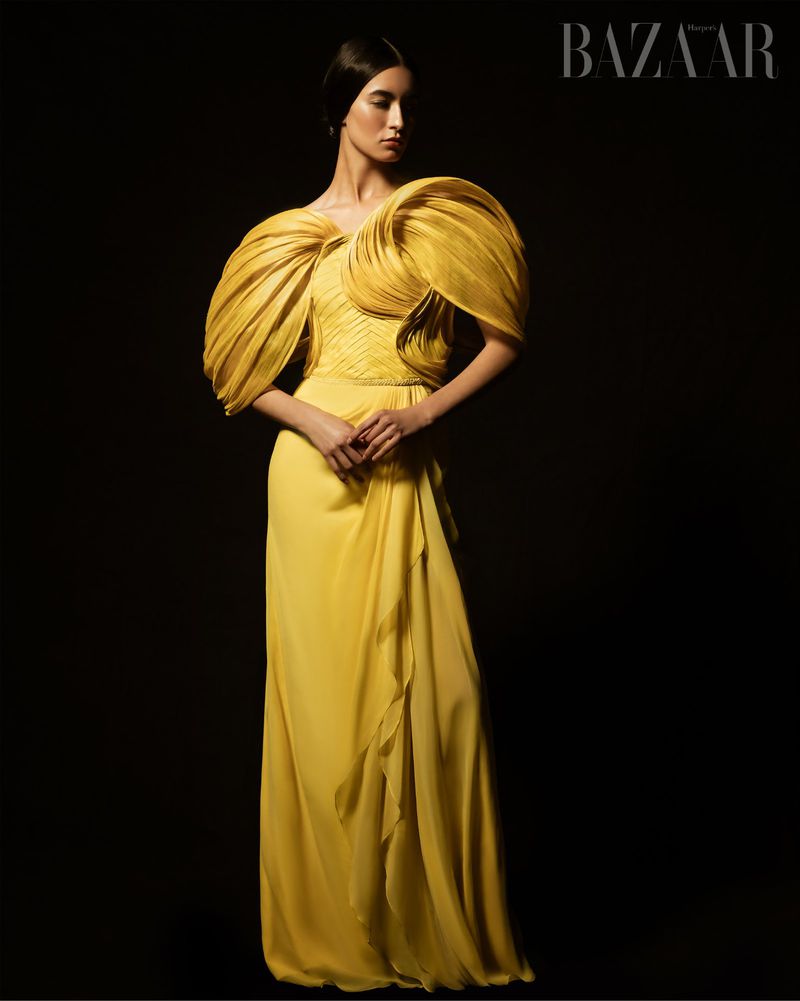 A woman in an elegant yellow gown with sculptural details poses in a fashion shot against a dark background for Bazaar Magazine.