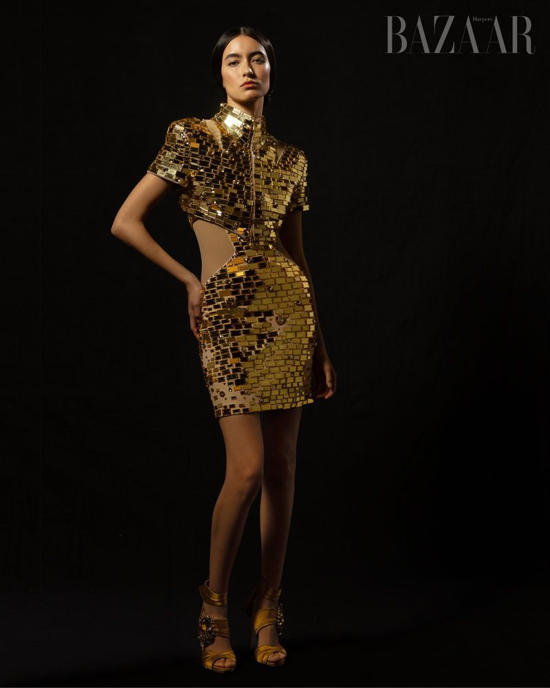 A fashion model poses in a gold, short, mirrored dress against a black background, her hands resting on her hip, with text visible.