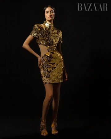 A fashion model poses in a gold, short, mirrored dress against a black background, her hands resting on her hip, with text visible.