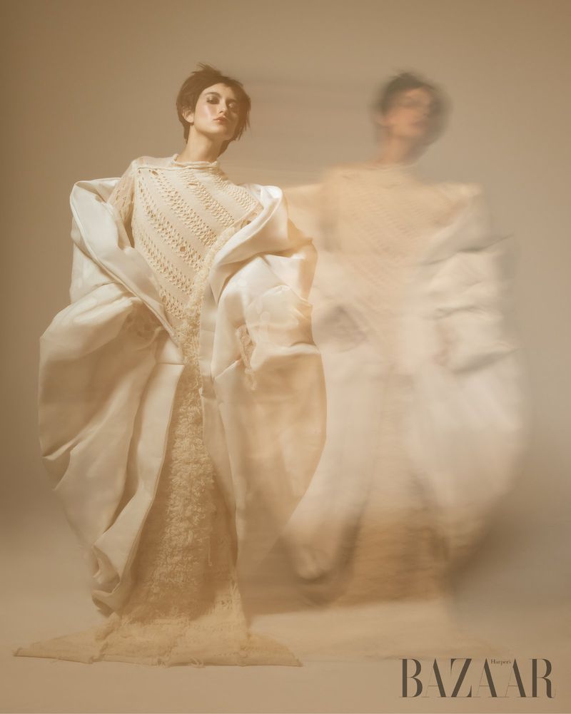 A woman is posing in a creamy white dress and jacket in a studio with motion blur and a creamy background, shot for Bazaar Magazine.