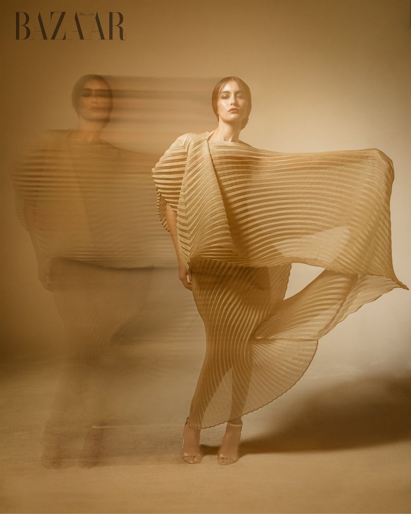 Fashion editorial shot of a woman in a flowing pleated dress with a motion-blurred double exposure, against a neutral background.