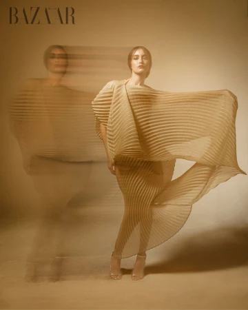Fashion editorial shot of a woman in a flowing pleated dress with a motion-blurred double exposure, against a neutral background.