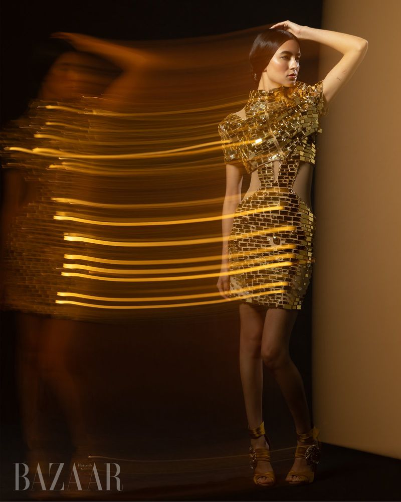 Fashion editorial shot of a woman in a gold sequin dress, posing with long exposure light streaks and dark background, in a studio.