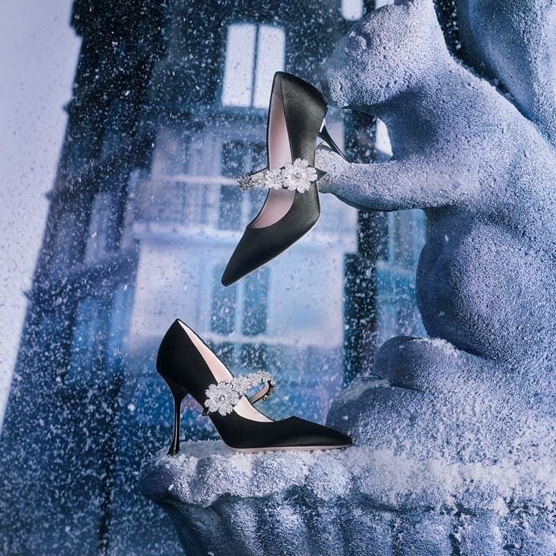 Elegant black high-heeled shoes with floral embellishments are displayed on a squirrel statue against a blurred architectural background.