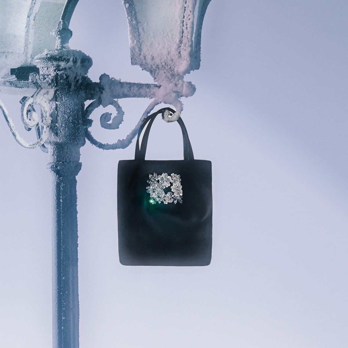 Elegant black purse adorned with a jewel flower, hanging from a snow-covered vintage lamppost against a bright blue-white sky.