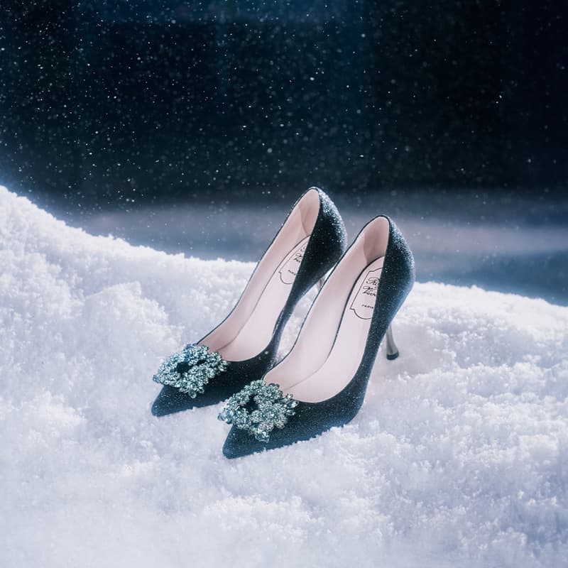 Elegant black high heel shoes adorned with glittering brooches, placed on a bed of snow against a dark, snowy backdrop. 