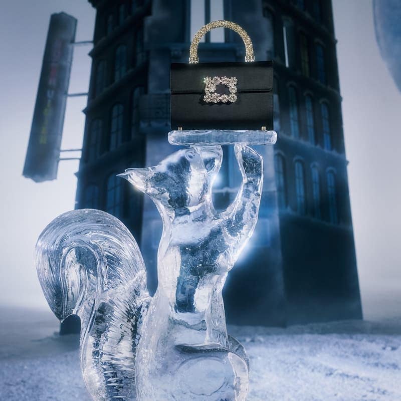 Black satin handbag with a jewel-encrusted handle and clasp rests on an ice sculpture of a squirrel against a snowy urban backdrop.