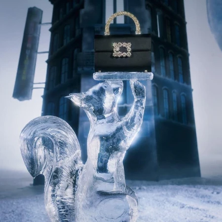 Black satin handbag with a jewel-encrusted handle and clasp rests on an ice sculpture of a squirrel against a snowy urban backdrop.