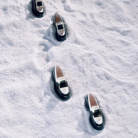 Four pairs of loafers, in a stepped formation, are on display in a studio setting with bright lighting and a snow-covered surface.