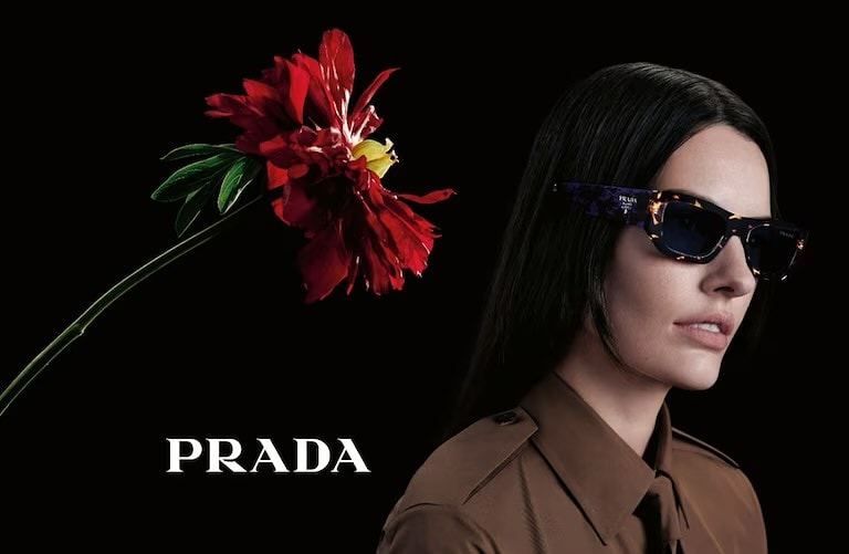 Portrait of a woman wearing sunglasses and a brown collared shirt, positioned near a large red flower against a black background.