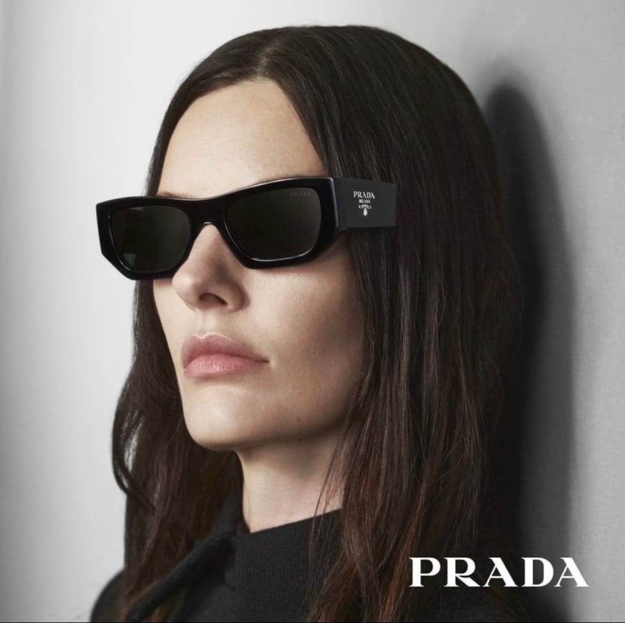 Brunette woman wearing black rectangular Prada sunglasses with logo in a studio shot for fashion, beauty, and celebrity brand promotion.