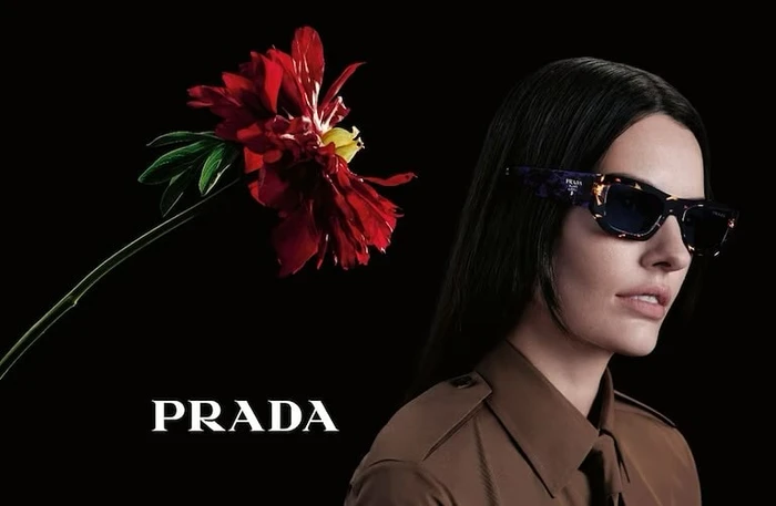 Model wearing dark sunglasses and brown shirt beside a large red flower against a black background for Prada Fall/Winter 2023 campaign.