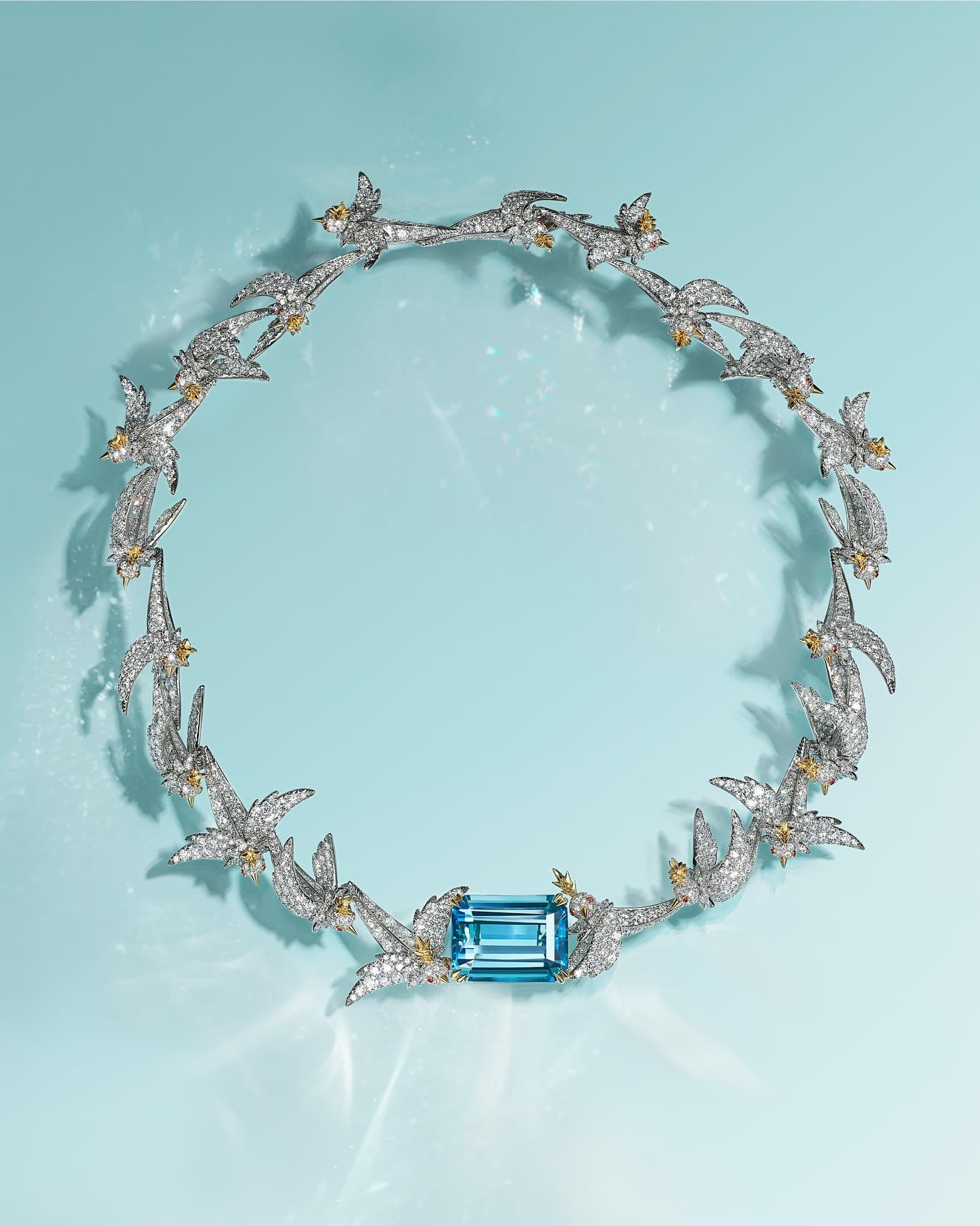 Elegant necklace featuring a circle of bird-shaped settings with diamonds and a large aquamarine stone against a light blue backdrop.