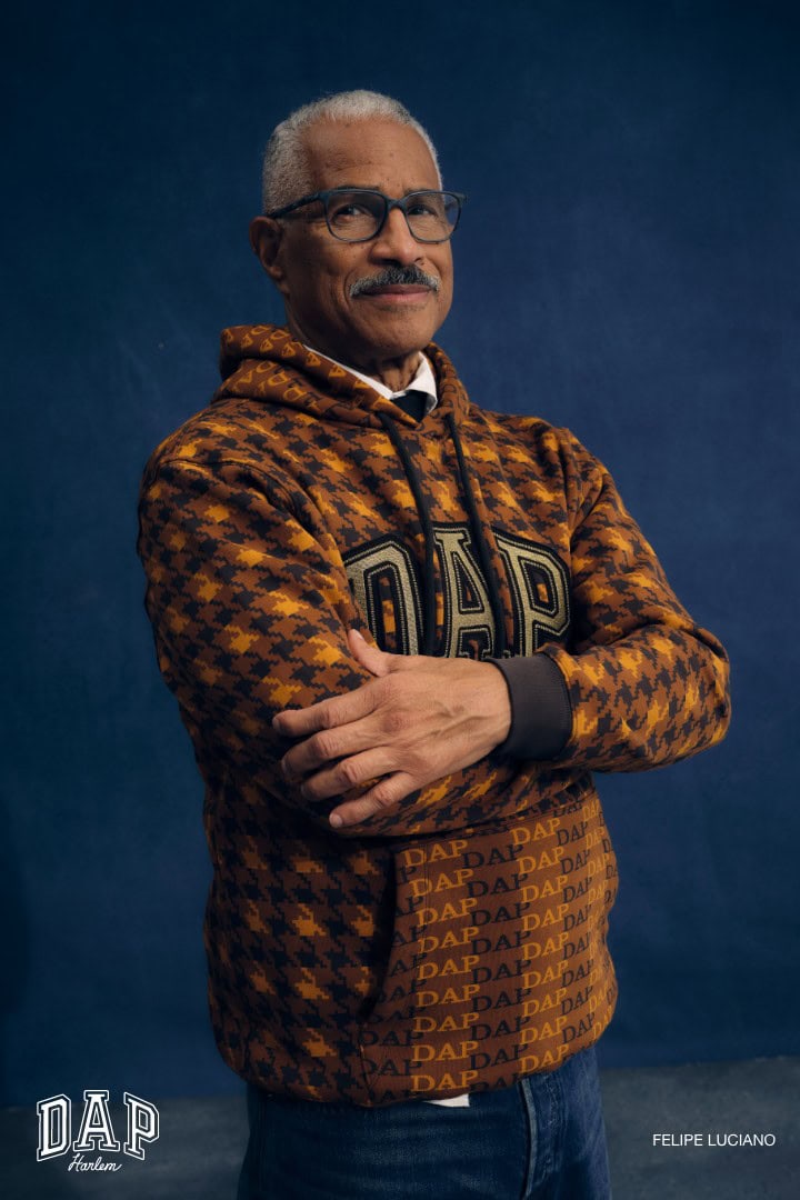 Portrait of mature adult man in patterned hoodie, glasses, and jeans against a dark blue background in a medium eye-level studio shot.