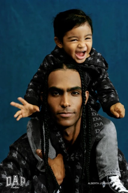 A man with braids carries a toddler on his shoulders; both wear matching patterned hoodies against a dark blue studio backdrop.