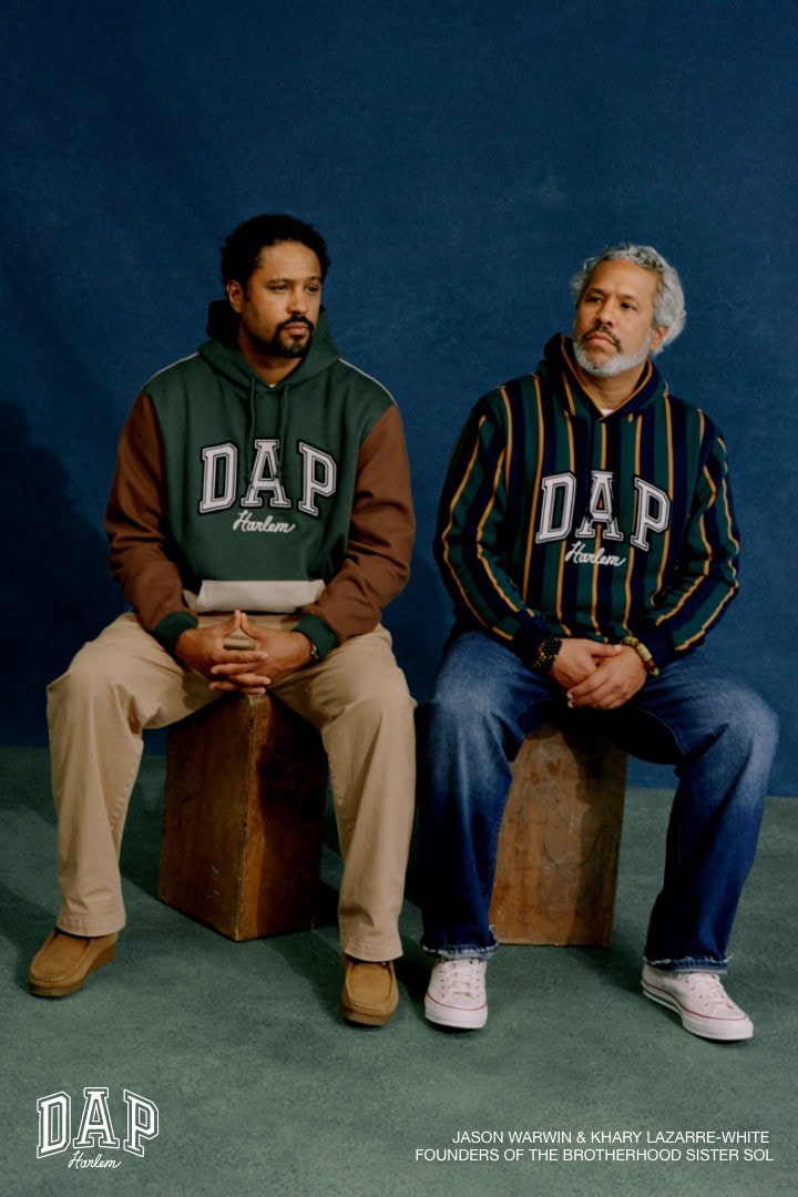 Two adult men sit side-by-side in sweatshirts, pants, and sneakers in a studio setting against a dark blue backdrop. Fashion.