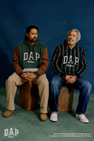 Two adult men sit side-by-side in sweatshirts, pants, and sneakers in a studio setting against a dark blue backdrop. Fashion.