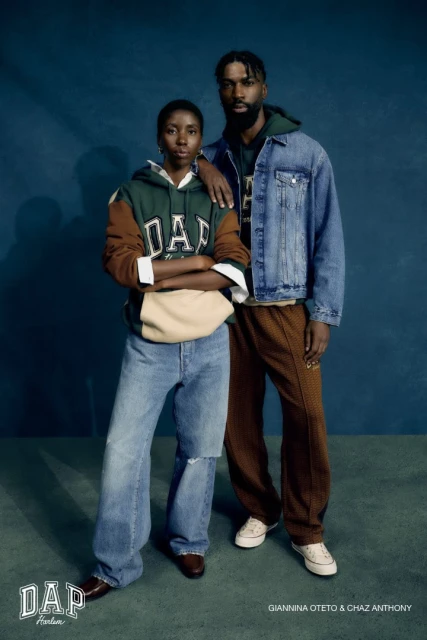 Two Black fashion models wearing vintage GAP clothing pose in a portrait, standing indoors in front of a dark teal backdrop.