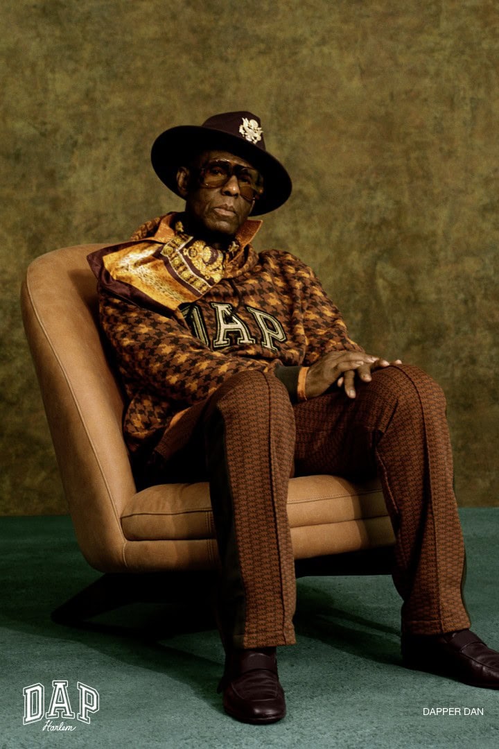 Adult male sits in a tan chair, wearing sunglasses, a patterned brown suit, and a wide-brim hat, against a mottled green background.