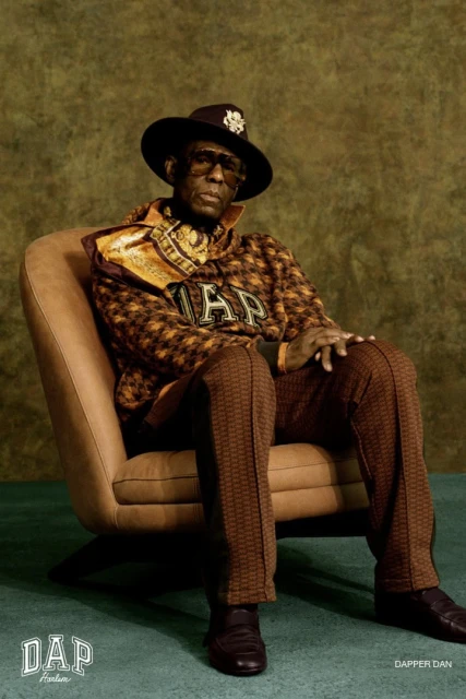 Adult male sits in a tan chair, wearing sunglasses, a patterned brown suit, and a wide-brim hat, against a mottled green background.