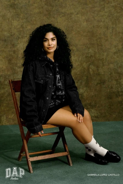 Portrait of a woman with curly hair wearing a denim jacket sitting on a wooden chair against an olive green backdrop.