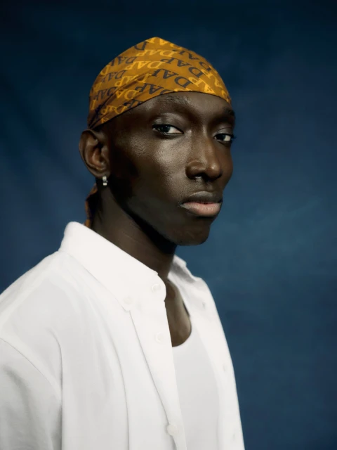 Portrait of a dark-skinned man with a patterned headscarf, wearing a white button-down shirt against a dark blue background in a studio shot.