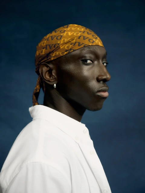 Portrait of a Black man in a white collared shirt and patterned headscarf against a mottled blue background in a studio setting.