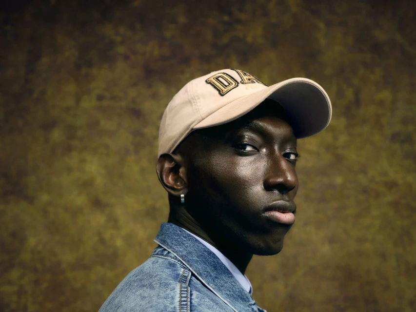 Portrait of a young Black man wearing a baseball cap and denim jacket, posing against a mottled brown and gold background.