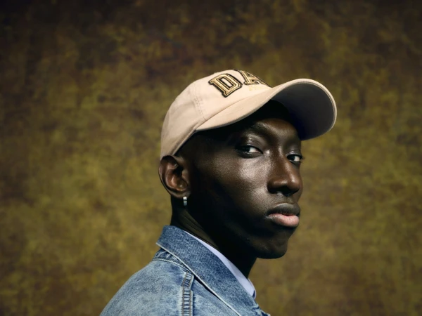 Portrait of a young Black man wearing a baseball cap and denim jacket, posing against a mottled brown and gold background.