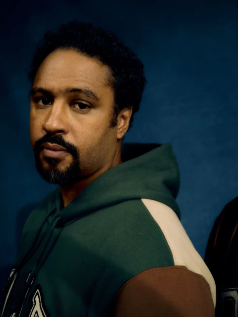 Portrait of a man with a goatee wearing a hooded sweatshirt against a deep blue background in a softly lit indoor setting.