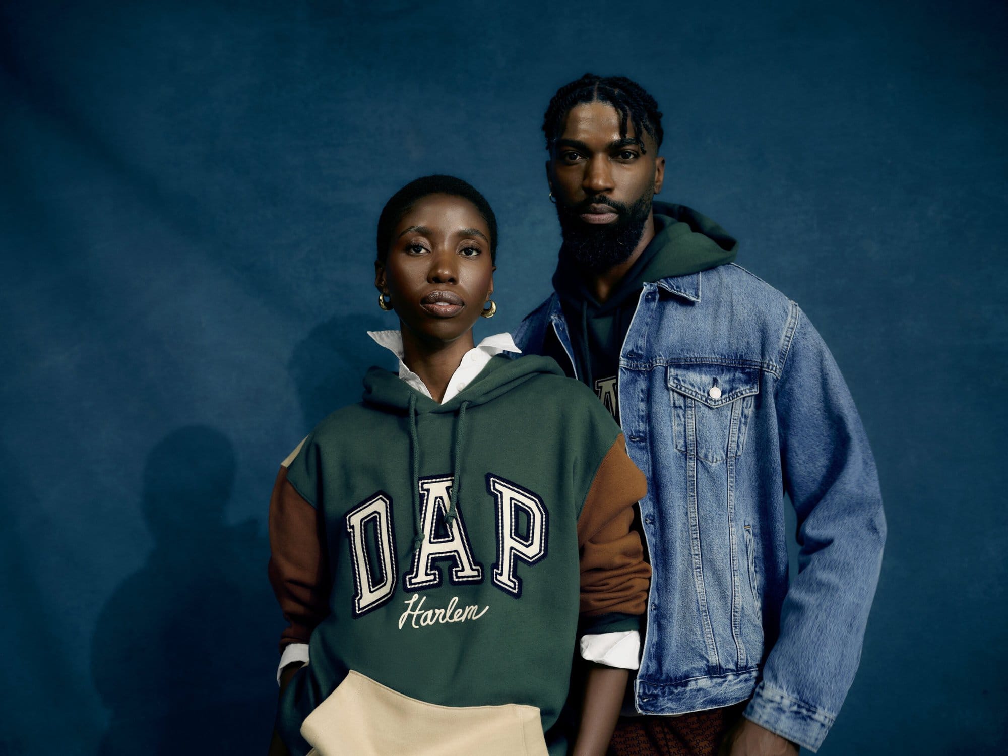 Two people pose in front of a blue wall. A woman wears a green and brown Gap hoodie, and a man wears a denim jacket and a green hoodie.