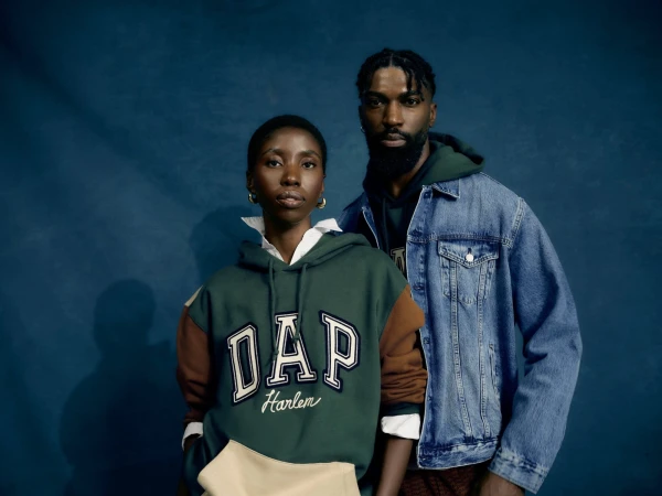 Two people pose in front of a blue wall. A woman wears a green and brown Gap hoodie, and a man wears a denim jacket and a green hoodie.