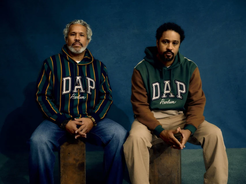 Two adult males are seated indoors against a blue backdrop. They are wearing matching DAP Harlem hooded sweatshirts in a medium shot.