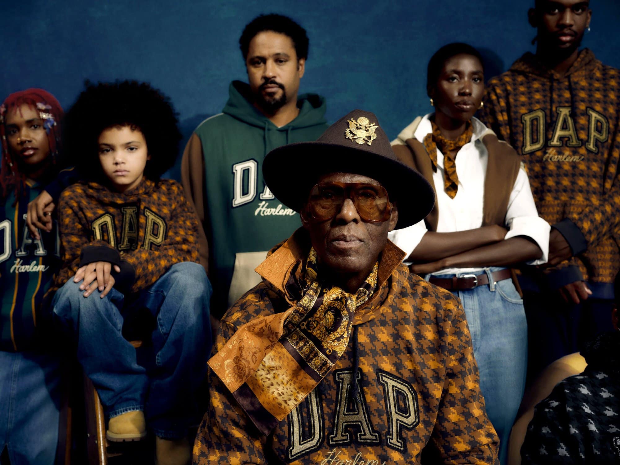 A diverse group of people wearing DAP Harlem hoodies are posing against a blue backdrop. The composition is a medium shot, highlighting fashion.