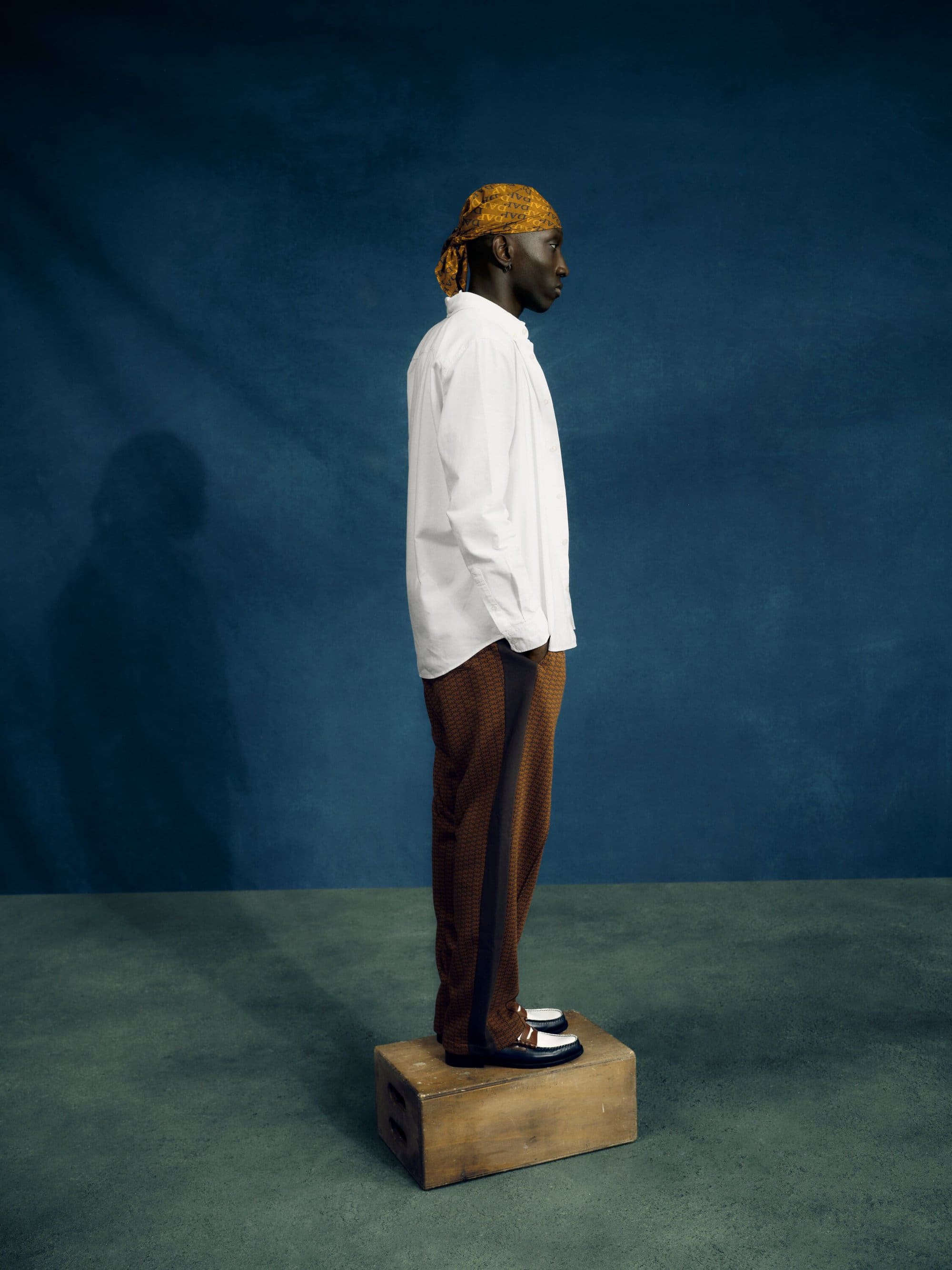 Dark-skinned adult male stands on a wooden box in a studio, wearing a white shirt, patterned brown pants, loafers, and a patterned head covering.