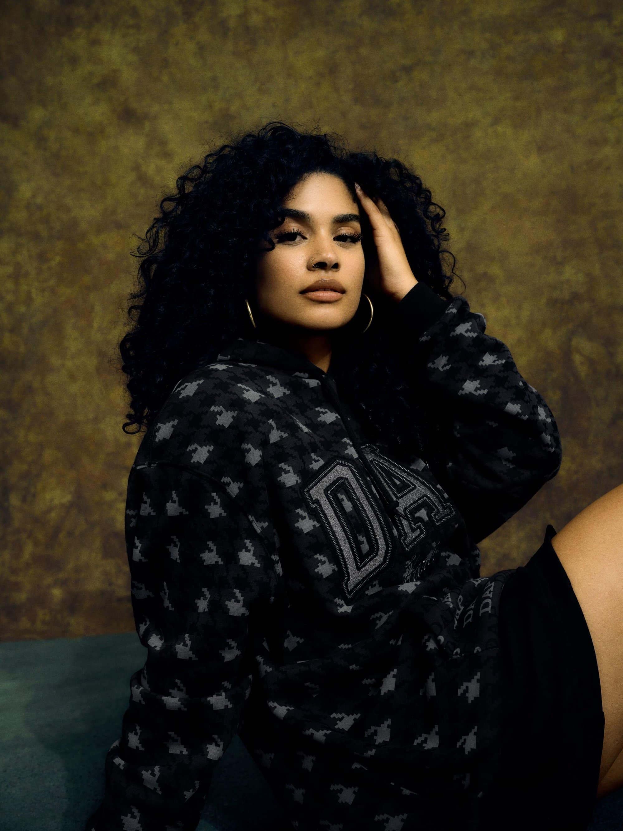 Portrait of a woman with curly hair wearing a patterned hoodie, seated against a mottled brown backdrop and teal velvet surface.