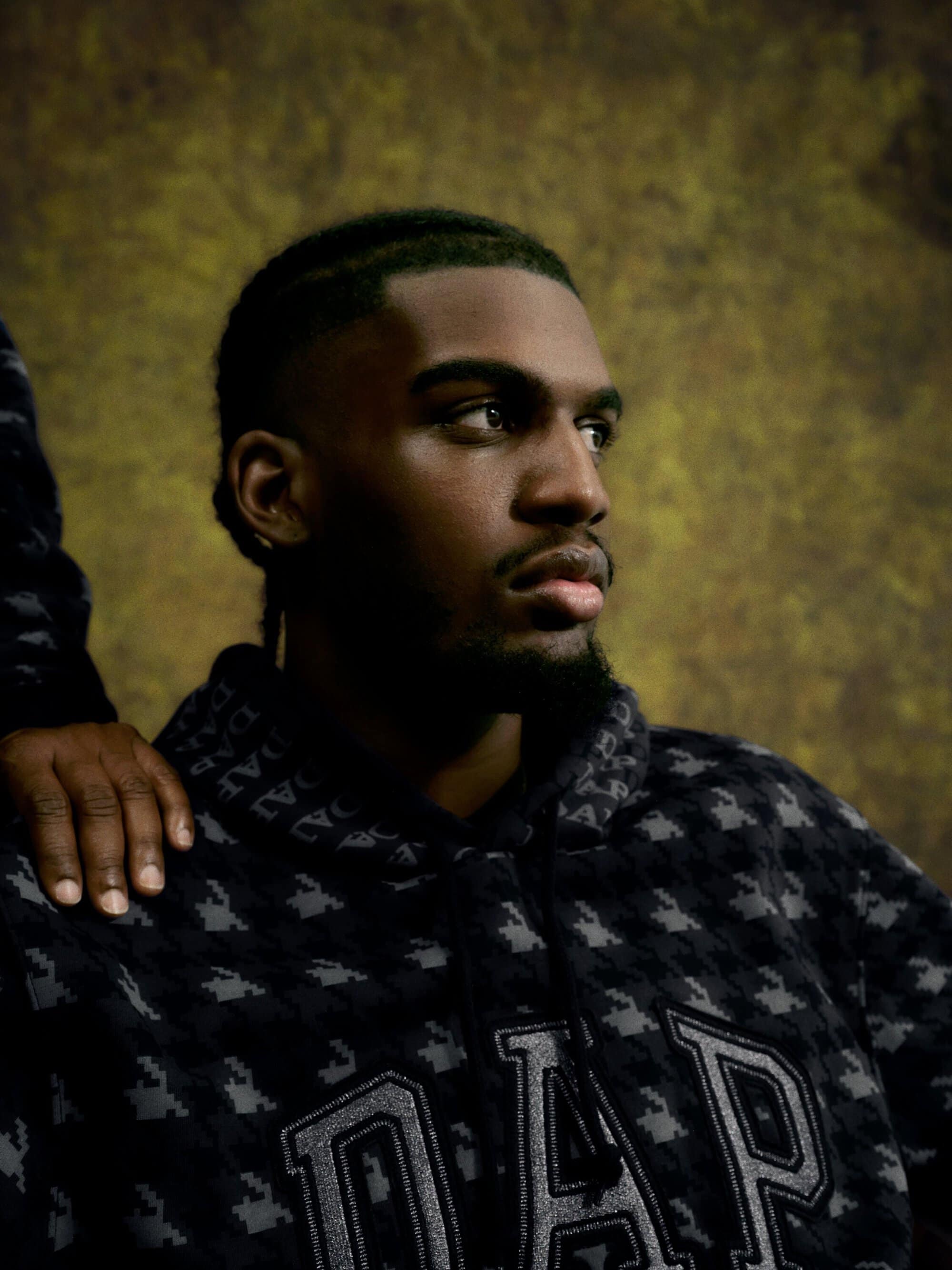 Portrait of a black male in patterned Gap hoodie with hand of second person on shoulder, against mottled backdrop in studio lighting.