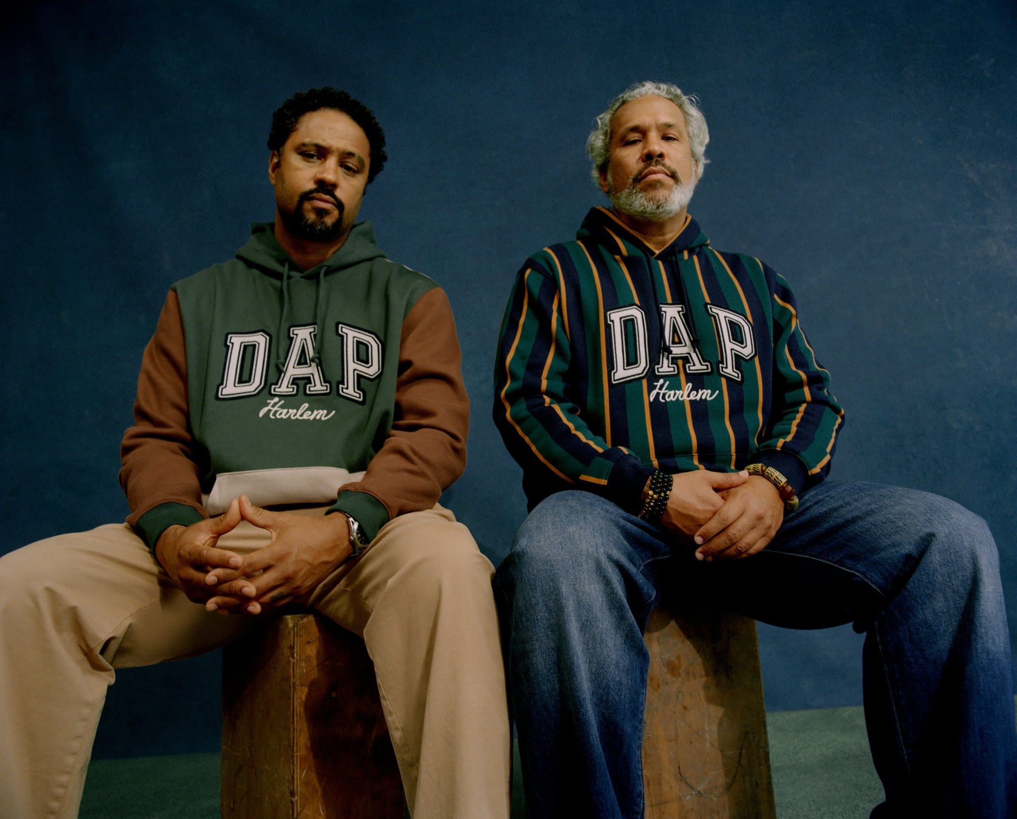 Two men seated indoors wearing DAP Harlem sweatshirts. One in green and brown, the other in blue striped, posing against a dark background.