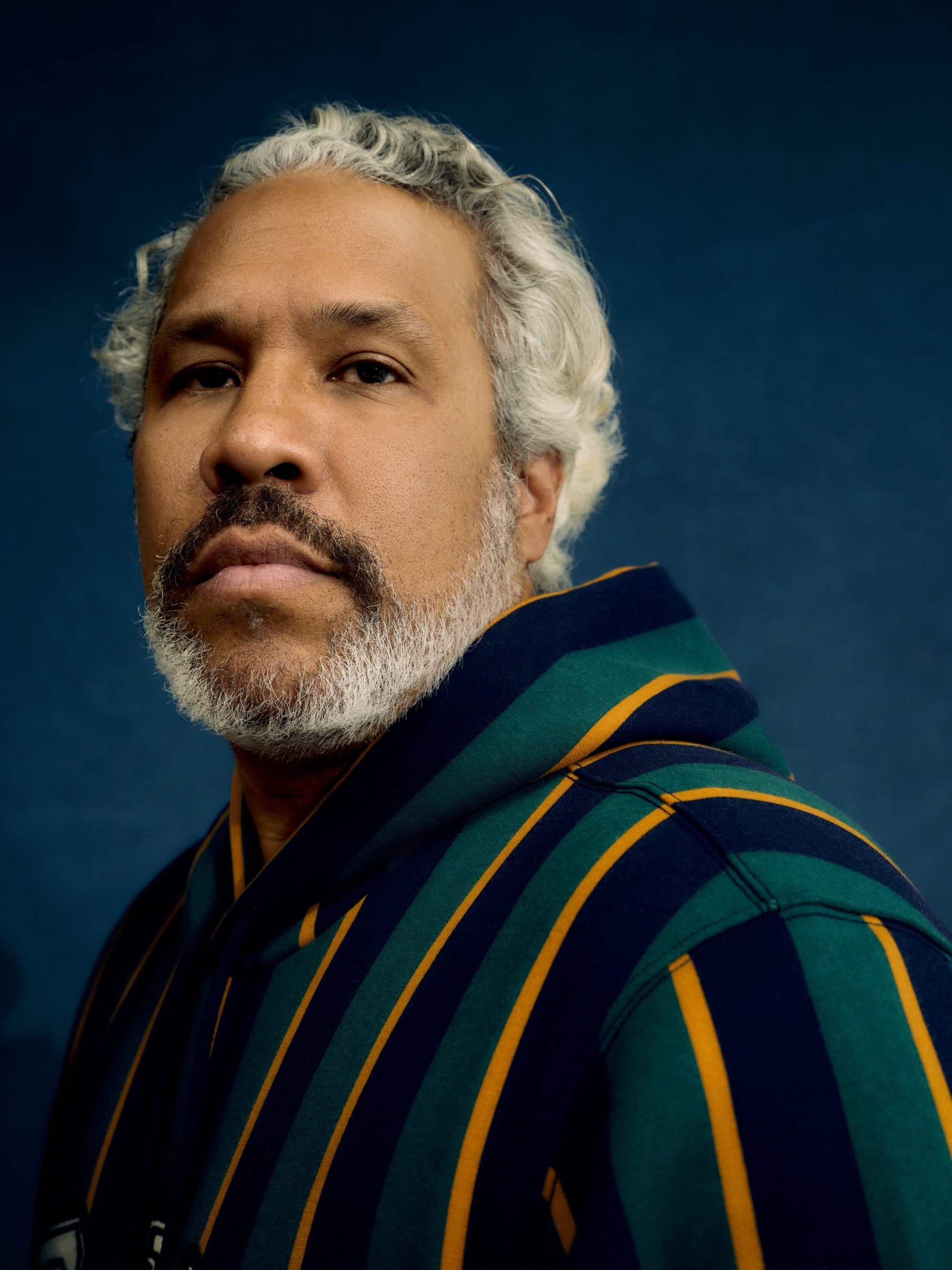 Portrait of a man with gray hair and a beard wearing a striped hoodie against a blue background, looking directly at the camera.