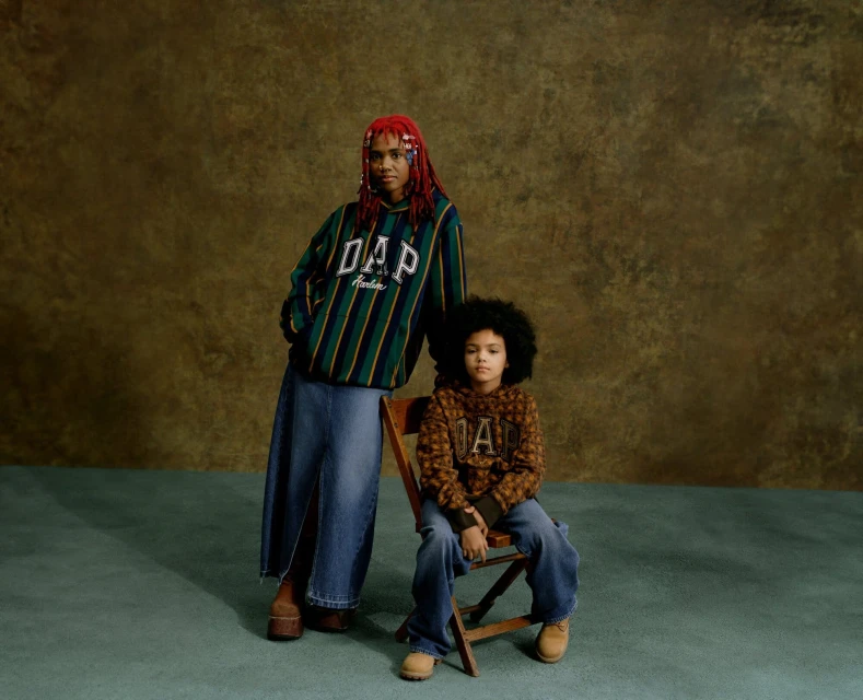 Adult woman and child pose indoors against a mottled brown backdrop wearing GAP-branded tops and denim bottoms in a fashion shot.
