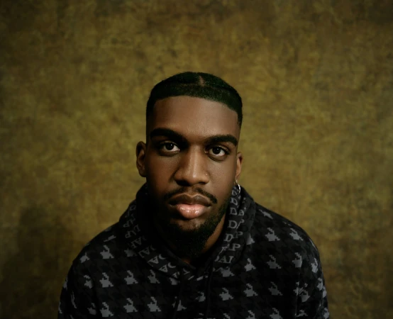 Portrait of a black man wearing a dark patterned hoodie against a textured olive-green backdrop for the Dap Gap Holiday 2023 campaign.