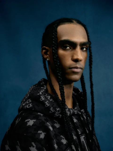 Close-up portrait of an adult Black male with braided hair wearing a patterned hoodie against a solid dark blue background.