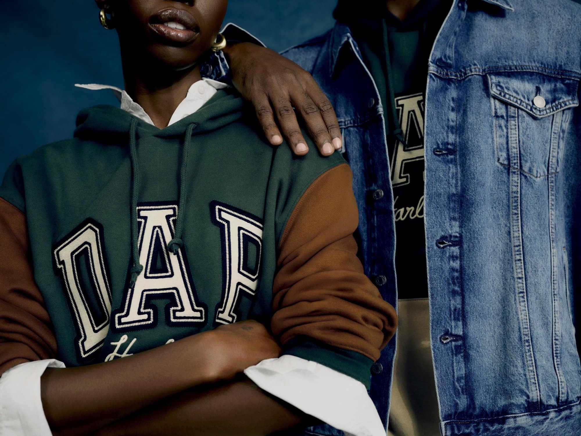 Close-up of two individuals posing together wearing Gap brand logo hoodies and a denim jacket against a deep blue background.